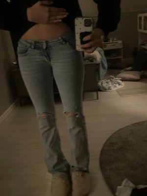 Ljusblå bootcut jeans  - Säljer ett par ljusblå bootcut jeans med hål över knäna och en rosa design på bakfickorna. Jeansen har låg midja och är ifrån Nelly i storlek S 