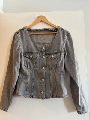 Grå jeansjacka med rundad urringning - Snygg grå jeans skjorta  med lång ärm och unik rundad urringning. Jackan har två bröstfickor med lock och knappar framtill. Figursydd modell som ger en modern siluett. Perfekt för dig som vill ha en lite annorlunda jeansjacka.
