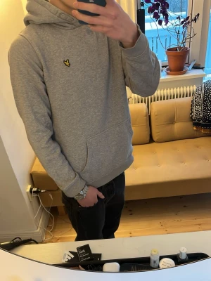 Grå hoodie från Lyle & Scott - Säljer en stilren grå hoodie från Lyle & Scott med klassisk gul logga på bröstet. Hoodien har huva, magficka och ribbade muddar vid ärmar och nederkant. Perfekt för en chill och avslappnad look. Materialet är mjukt och skönt bomullstyg.