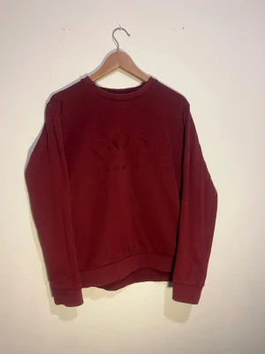 Vinröd sweatshirt från Hugo Boss - Snygg vinröd sweatshirt från Hugo Boss med broderad logga framtill. Tröjan har rund halsringning, långa ärmar och ribbade muddar vid ärmslut och nederkant. Tillverkad i mjukt material som känns skönt mot huden. Perfekt för en avslappnad och stilren look.