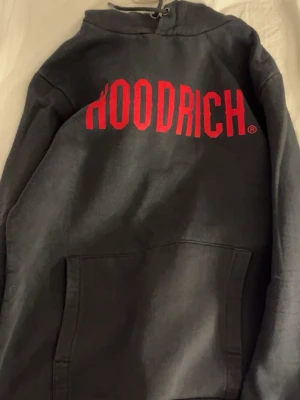Svart hoodie från Hoodrich med röd logga - Svart hoodie från Hoodrich med stor röd logga över bröstet. Klassisk känguruficka framtill och justerbar huva med snören. Perfekt för en streetwear-look och riktigt skön att bära till jeans eller joggers. M men mer S. Buda gärna!! 