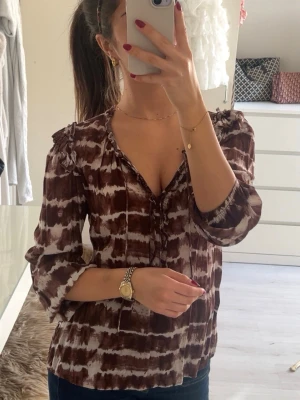 Brun och vit blus med volang - Säljer en brun och vit mönstrad blus med volangdetaljer vid axlarna och v-ringning. Blusen har trekvartsärm och ett avslappnat fall. Perfekt för dig som gillar boho-vibbar och vill ha något unikt i garderoben.