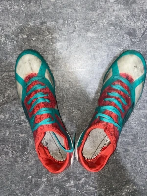 Färgglada Umbro fotbollsskor - Säljer ett par Umbro fotbollsskor i vitt, turkost och rött med mönstrad ovandel och turkosa snören. Skorna har låg profil och är designade för bra grepp på planen. Perfekta för dig som vill sticka ut på matchen!