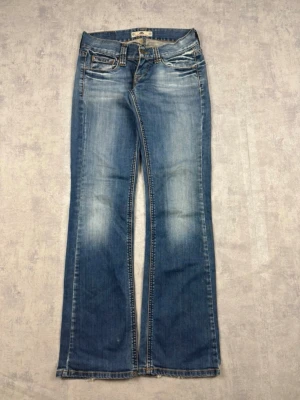 Blå bootcut jeans med slitningar - Säljer ett par blå bootcut jeans med snygga slitningar och ljusa partier framtill. Jeansen har klassisk femficksmodell, låg midja och utsvängda ben. Perfekta för dig som gillar retrovibbar och avslappnad stil.