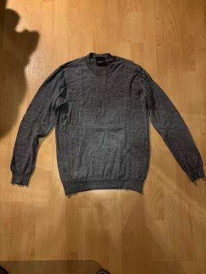 Grå stickad tröja Oscar Jacobson - Snygg grå stickad tröja från Oscar Jacobson i klassisk modell. Tröjan har rund halsringning, långa ärmar och ribbade muddar vid ärmslut och nederkant. Perfekt att matcha med jeans eller chinos för en clean look.