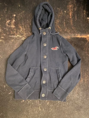 Mörkblå cardiganhoodie från Hollister - Mörkblå hoodie från Hollister med knappar framtill och två st fickor. Klassisk huva med dragsko och Hollisters logga i rött och vitt på bröstet. Tillverkad i mjukt sweatshirtmaterial, perfekt för en avslappnad stil. Sitter som XS men är funkar på S också! Skriv vid frågor☺️