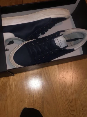 Marinblå sneakers från Armani Exchange - Snygga marinblå sneakers från Armani Exchange med vita detaljer och logga på hälen. Skorna har klassisk låg profil, svarta skosnören och vit sula. Perfekta för dig som gillar stilrena och moderna sneakers.