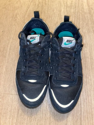 Svarta Nike sneakers med vit swoosh - Säljer ett par svarta Nike sneakers med vit swoosh på sidan och reflekterande detaljer framtill. Skorna har svart mesh och mockaliknande material, samt turkosa innersulor och svart snörning. Perfekta för dig som gillar stilrena och sportiga skor.