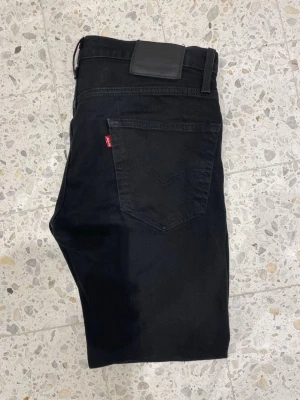 Svarta Levi's jeans med rak passform - Säljer ett par klassiska svarta jeans från Levi's 502 med rak passform och fem fickor. Jeansen har en diskret röd Levi's-tag på bakfickan och svarta sömmar för en clean look. Perfekta för dig som gillar tidlös och enkel stil.