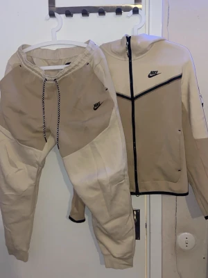 Beige Nike loungewear set - Säljer ett beige och ljusbrunt loungewear set från Nike med matchande hoodie och byxor. Setet har svarta detaljer, dragkedja på tröjan och snörning i midjan på byxorna. Båda delarna har Nike-logga broderad på bröstet och benet. Perfekt för chill dagar hemma. byxor storlek M tröja Storlek S