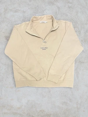 Beige halfzip hoodie Calvin Klein - Snygg beige hoodie från Calvin Klein Jeans med halv dragkedja framtill och broderad logga på bröstet. Tillverkad i mjukt bomullsmaterial och har ribbade muddar vid ärmslut och nederkant. Perfekt för en chill och stilren look.