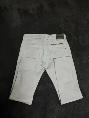 Vita jeans från Replay Jeans - Säljer ett par vita jeans från Replay Jeans med raka ben och coola detaljer som stora bakfickor och en dragkedja på ena fickan. Jeansen har bälteshällor och är tillverkade i ett slitstarkt bomullsmaterial. Perfekta för dig som vill sticka ut med en clean look.