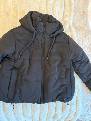 Svart pufferjacka  -  en svart pufferjacka från Design by Amor i storlek XS. Jackan har huva med snörning, dragkedja framtill och en oversized look. Perfekt för kalla dagar.