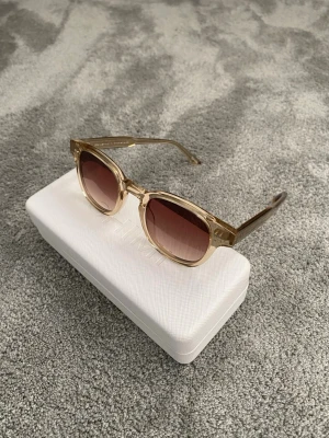 Solglasögon - Snygga solglasögon från CHIMI ECRU 01 med transparent beige båge och runda, bruna tonade glas. Klassisk och trendig design som passar till många stilar. Kommer med ett vitt hårt fodral för säker förvaring.