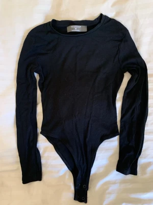 Svart långärmad body Primark XS - En enkel och stilren svart body från Primark i storlek XS. Bodyn har långa ärmar, rund halsringning och knäppning i grenen. Tillverkad i mjukt stretchigt material som sitter snyggt på kroppen. Perfekt att matcha med jeans eller kjol. Endast använd en gång!