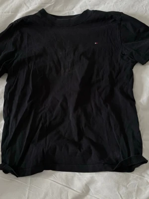 Svart t-shirt från Tommy Hilfiger - Snygg svart t-shirt från Tommy Hilfiger med liten diskret logga på bröstet. Klassisk rund halsringning och mjukt bomullsmaterial som känns skönt mot huden. Perfekt basplagg till garderoben och lätt att matcha med allt.
