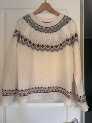 Vit stickad tröja med mönster - Säljer en vit stickad tröja med rund hals och färgglatt nordiskt mönster i blått, rött, svart och beige runt hals, ärmslut och nederkant. Tröjan har långa ärmar och ribbade muddar. Perfekt för dig som gillar klassisk och mysig stil.