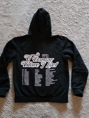 Svart hoodie Hov1 Vi Dansar Vidare - Svart hoodie från Hov1 med stor vit text på ryggen 'Vi Dansar Vidare I Livet' och turnédatum. Klassisk huva och muddar vid ärmar och midja. Perfekt för dig som gillar svensk musik och vill ha en unik merch-stil.