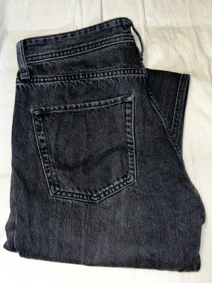 Svarta raka jeans från Jack & Jones - Säljer ett par svarta jeans från Jack & Jones  med rak passform och klassisk femficksdesign. Jeansen har en tvättad look och är tillverkade i denim, vilket ger en avslappnad och trendig stil. Stl W30 L32