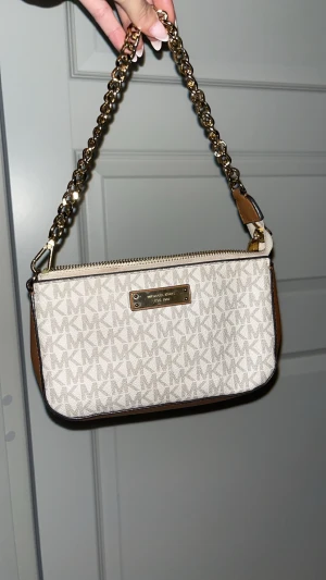 Beige axelremsväska Michael Kors - Snygg beige handväska från Michael Kors med MK-logga över hela väskan. Väskan har guldfärgad kedjerem och detaljer i brunt läder. Praktisk dragkedja upptill och metallskylt framtill. Perfekt storlek för det viktigaste och ger en lyxig känsla.