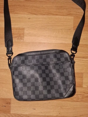 Louis Vuitton axelväska i damier-mönster - Snygg axelväska från Louis Vuitton i klassiskt svart och grått damier-mönster. Väskan har justerbar axelrem i textil och detaljer i svart läder. Perfekt storlek för det viktigaste och en stilren look med präglad logga på hörnet.