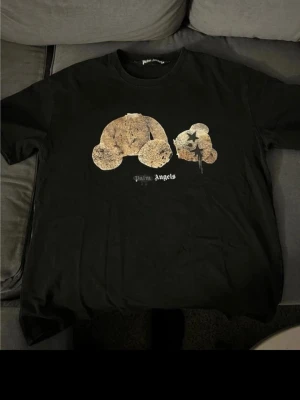 Svart Palm Angels t-shirt med björn - Säljer en svart t-shirt från Palm Angels med ikoniskt björnmotiv på bröstet och logga under. T-shirten har rund hals och korta ärmar. Perfekt för dig som gillar streetwear och vill sticka ut med en unik design.
