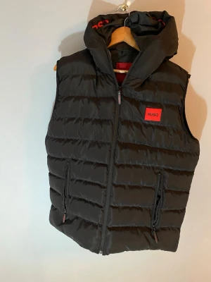 Svart dunväst från HUGO med huva - Snygg svart dunväst från HUGO med avtagbar huva och röda detaljer. Västen har två dragkedjeförsedda fickor och en röd HUGO-logga på bröstet. Perfekt för lager-på-lager och streetwear-stil.