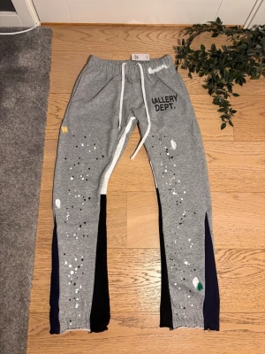 Grå mjukisbyxor Gallery Dept. - Grå mjukisbyxor från Gallery Dept. med vita och svarta färgstänk, tryckt logga på vänster sida och kontrastpaneler i mörkblått längs benen. Byxorna har resår i midjan med vit dragsko och muddar vid bensluten. Perfekt för en avslappnad och trendig look.