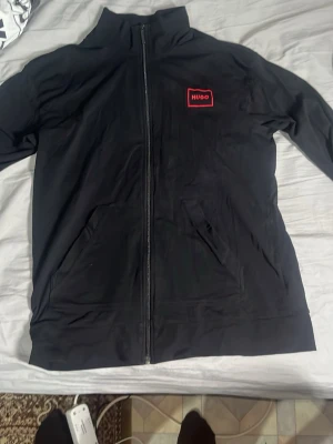 Svart zip hoodie från Hugo - Snygg svart hoodie från Hugo med dragkedja framtill och två fickor. Diskret röd Hugo-logga på bröstet och matchande röd etikett på insidan. Tillverkad i mjukt material som känns skönt mot huden. Perfekt för en avslappnad och stilren look om ni inte gillar det satta priset så går det att förhandla.