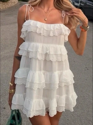 Vit volangklänning med spetsdetaljer - Supersöt vit kortklänning med flera lager volanger och spetsdetaljer. Axelbandslös look med tunna knytband på axlarna. Luftigt material som känns lätt och bekvämt.🌸perfekt till studenten!