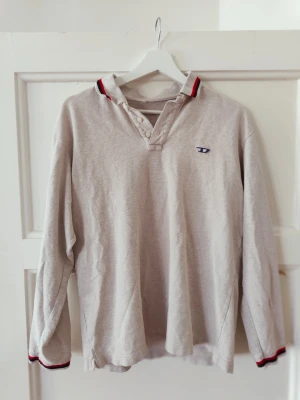 Diesel piké - Klassisk grå långärmad polotopp från Diesel med knappar vid halsen och diskret logga på bröstet. Kragen har röda och svarta detaljer som ger en sportig touch. Perfekt till jeans eller chinos.