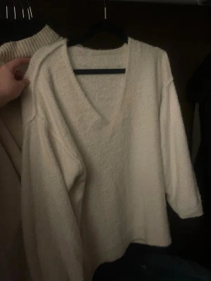 Krämvit stickad tröja med v-ringning - Mysig krämvit stickad tröja med djup v-ringning och avslappnad passform. Tröjan har långa ärmar och är gjord i ett mjukt, fluffigt material som känns skönt mot huden. Perfekt för lager på lager och en chill look.