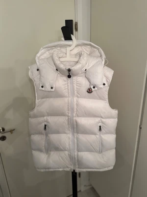 Vit dunväst från Moncler med huva - Snygg vit dunväst från Moncler med avtagbar huva och två dragkedjefickor framtill. Västen har en puffig design och klassisk Moncler-logga på bröstet. 