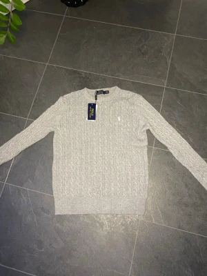 Ljusgrå kabelstickad tröja Polo Ralph Lauren - Snygg ljusgrå kabelstickad tröja från Polo Ralph Lauren med rund halsringning och diskret broderad logga på bröstet. Tröjan har långa ärmar och ribbade muddar vid ärmslut och nederkant. Perfekt för lager på lager och klassisk stil.