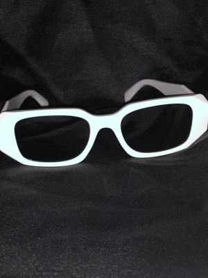 Vita glasögon från Prada - Snygga vita solglasögon från Prada med breda skalmar och svarta glas. Tillverkade i Italien och har en modern, kantig form som ger en cool och trendig look. Perfekta för dig som vill sticka ut med en exklusiv accessoar.