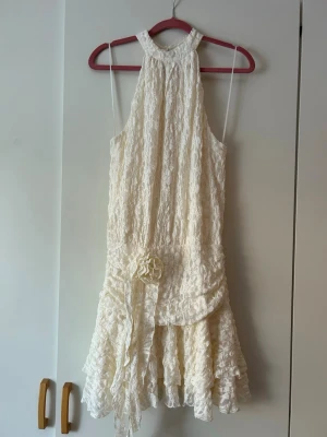 Vit spetsklänning med volang - Supersöt vit klänning i spets med volangkant och dekorativ blomdetalj i midjan. Klänningen är ärmlös, har halterneck och en asymmetrisk nederdel som ger en romantisk vibe. Perfekt för sommarens alla tillfällen! Inte använt än.