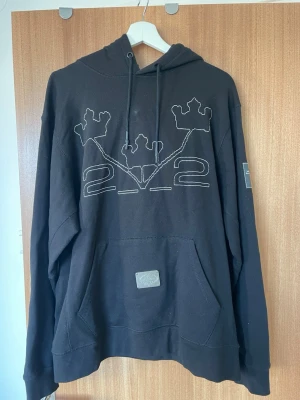 Svart hoodie Stockholm Beauty Group - Svart hoodie med stor magficka och huva, dekorerad med broderade kronor och siffrorna 22 på bröstet. Metallbricka med Stockholm Beauty Group-logga på fickan. Justerbar dragsko i huvan och patch på ärmen. Observera att ena ”ringen” vid kragen till snöret lossnat samt lite av broderingen gått upp, se bilder. 