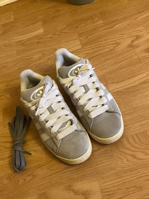 Grå Adidas Campus sneakers - Säljer ett par grå Adidas Campus sneakers i mocka med vita klassiska stripes på sidorna och vit sula. Skorna har låga skaft och kommer med extra grå skosnören. Perfekta för dig som gillar stilrena och tidlösa sneakers. Har knappt använt de, jag säljer de också för att jag vill ha en annan färg istället. (De är helt rena) 😊