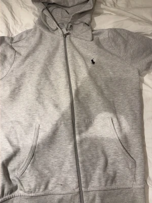Grå hoodie från Polo Ralph Lauren - Snygg ljusgrå hoodie från Polo Ralph Lauren med dragkedja framtill, korta ärmar och två fickor. Klassisk broderad logga på bröstet och justerbar huva med snören. Tillverkad i mjukt bomullsmaterial som känns skönt mot huden.