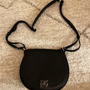 Svart axelväska från DKNY - Snygg svart axelväska från DKNY i skinn. Väskan har en justerbar axelrem och ett stilrent design med DKNY-logga framtill. Perfekt för att bära dina nödvändigheter med stil. Den är helt ny och aldrig använd. Nypris ligger på 3500kr.