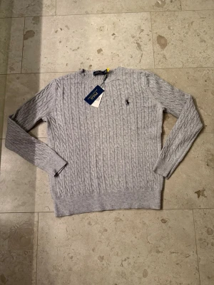 Grå kabelstickad tröja från Polo Ralph Lauren - Snygg grå kabelstickad tröja från Polo Ralph Lauren med klassisk rund halsringning och broderad logga på bröstet. Tröjan har långa ärmar och ribbade muddar vid ärmslut och nederkant. Helt ny med lappar på som ni kan se.