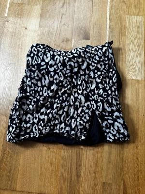 Kort leopardmönstrad kjol - Snygg kort kjol med leopardmönster i svart, beige och grå toner. Kjolen har en liten slits på sidan för extra edge och är tillverkad i ett mjukt, följsamt material. Perfekt för dig som vill sticka ut med en trendig look.