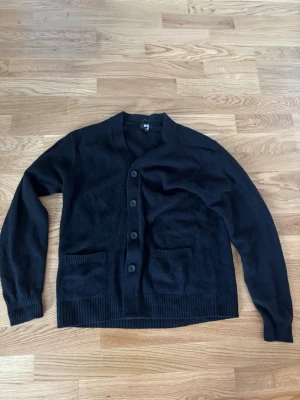 Svart kofta/cardigan  från Uniqlo, S - Klassisk svart kofta från Uniqlo med v-ringning och knäppning framtill. Två fickor fram och ribbade muddar vid ärmslut och nederkant. Perfekt att slänga över en t-shirt eller skjorta för en clean look.