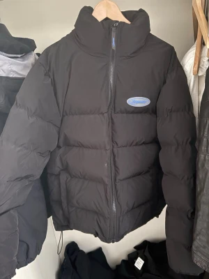 Svart pufferjacka från Trapstar - Säljer en svart pufferjacka från Trapstar med blå logga på bröstet och stor broderad Trapstar-text i blått på ryggen. Jackan har hög krage, dragkedja framtill och en klassisk oversized puffer-look. Perfekt för kalla vinterdagar och riktigt street.