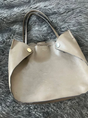 Beige handväska från Guess - Stilren beige handväska från Guess med två smala axelband och silverfärgade tryckknappar. Väskan har en mjuk, slät yta i konstläder och en enkel, rymlig design. Perfekt för dig som gillar minimalistisk stil och vill ha plats för allt du behöver.