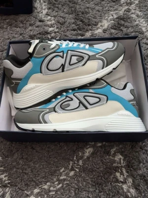 CD grå/blå sneakers från Dior - Säljer ett par snygga Dior sneakers med stora CD-loggor på sidan. Skorna har en mix av grått, blått, vitt och beige med mesh och läderdetaljer. Tjock vit sula och vita skosnören ger en modern vibe. Perfekta för dig som gillar statement sneakers.