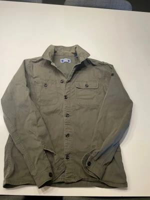 Olivgrön overshirt från Jack & Jones - Stilren olivgrön overshirt från jack & jones med två bröstfickor med lock och knappar. Skjortjackan har klassisk krage, svarta knappar framtill och på ärmsluten samt diskret logotyp på ärmen. Tillverkad i ett slitstarkt bomullstyg som ger en avslappnad look.  (Skriv om ni är intresserade ☺️)