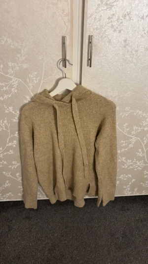 Beige stickad hoodie med huva - Säljer en beige stickad hoodie med huva och dragsko. Tröjan har långa ärmar och ribbade muddar vid ärmslut och nederkant. Perfekt för en avslappnad och stilren look. Materialet känns mjukt och bekvämt mot huden.