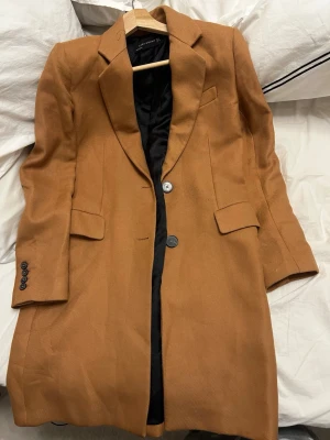 Kamelbrun kappa från Zara Woman - Stilren kamelbrun kappa från Zara Woman med klassisk krage, två knappar. Kappan har två fickor med lock och dekorativa knappar vid ärmsluten. Perfekt för dig som gillar en tidlös och elegant look.