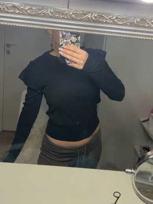 Svart topp med volangdetaljer - Säljer en svart långärmad topp med volangdetaljer över axlarna. Toppen har en tight passform och är croppad så den slutar vid midjan. Perfekt att styla med både jeans och kjol för en trendig look.
